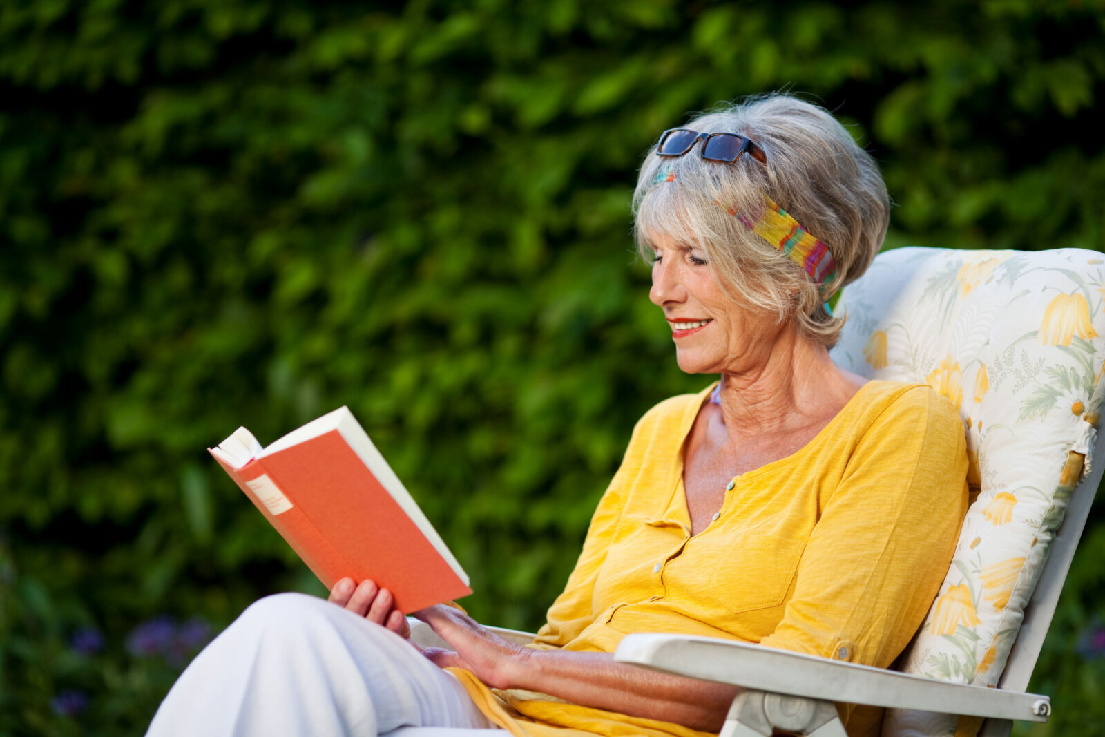 A senior woman is reading a book while sitting on a chair at the park with a smile on her face.
