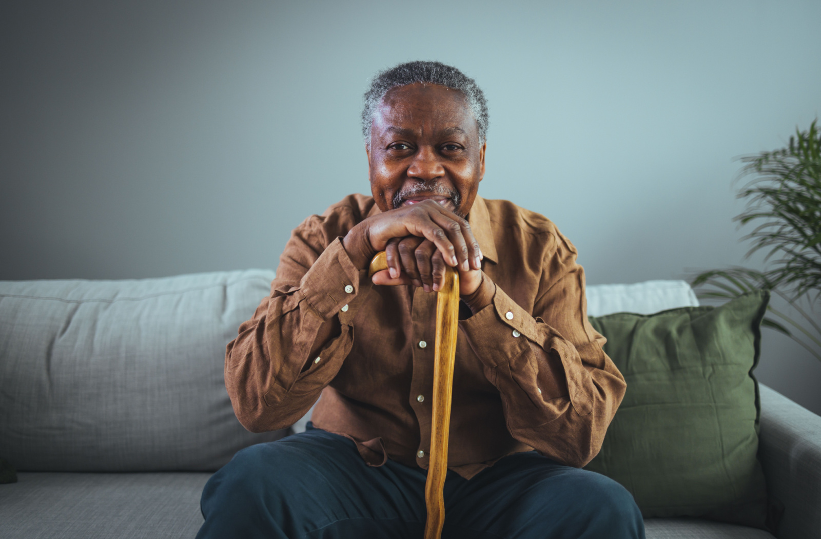 A senior man with short gray hair holding a cane and smiling while sitting on a sofa.