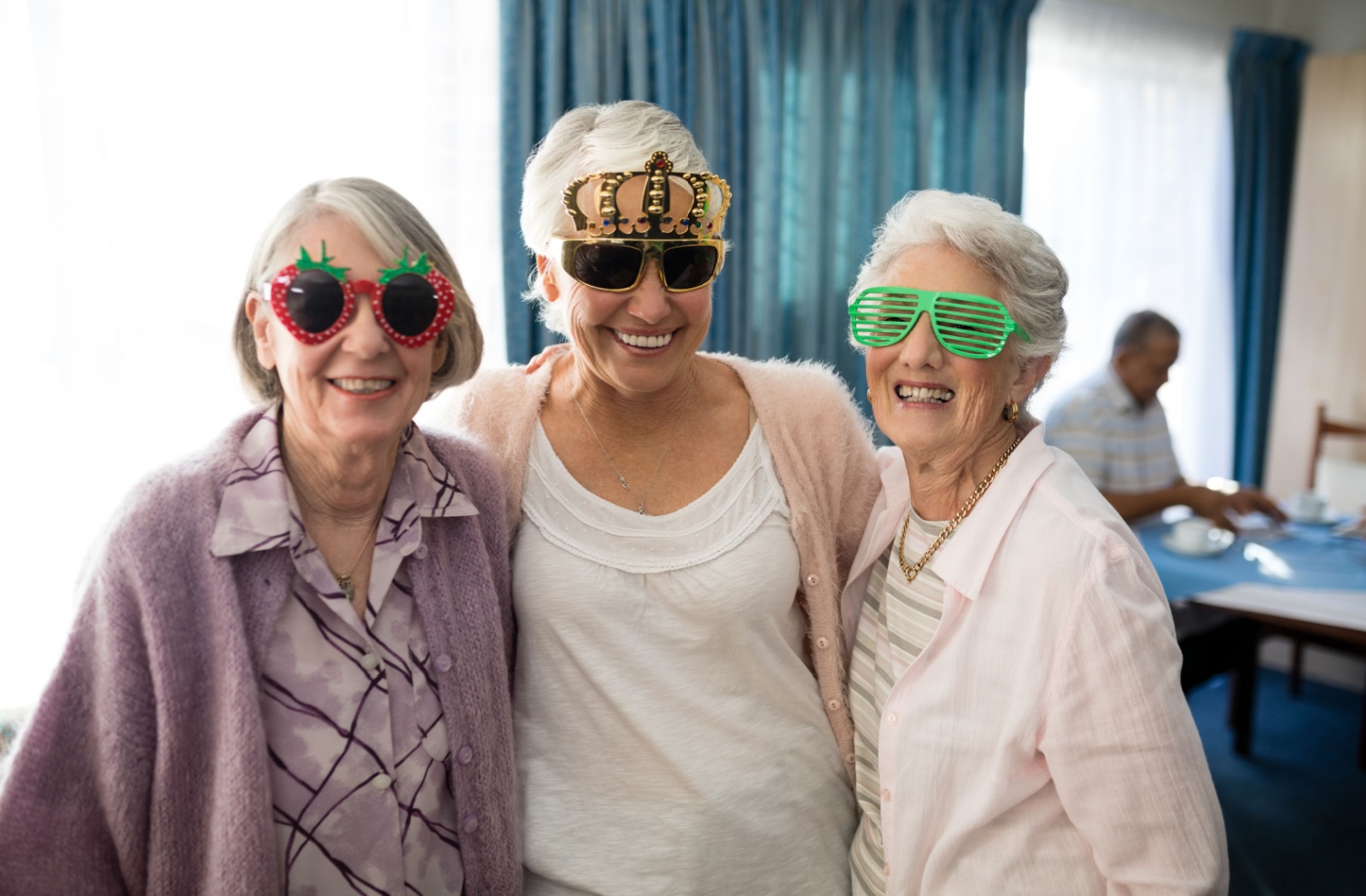 3 seniors are standing together in a well-lit dining room. They have their arms around each other and are smiling at the camera. Each person is wearing a pair of funny glasses. One is wearing strawberry shaped sunglasses, the second is wearing frames with a small, gold crown atop the frames, and the third is wearing bright green glasses with thin slits across the eyes.