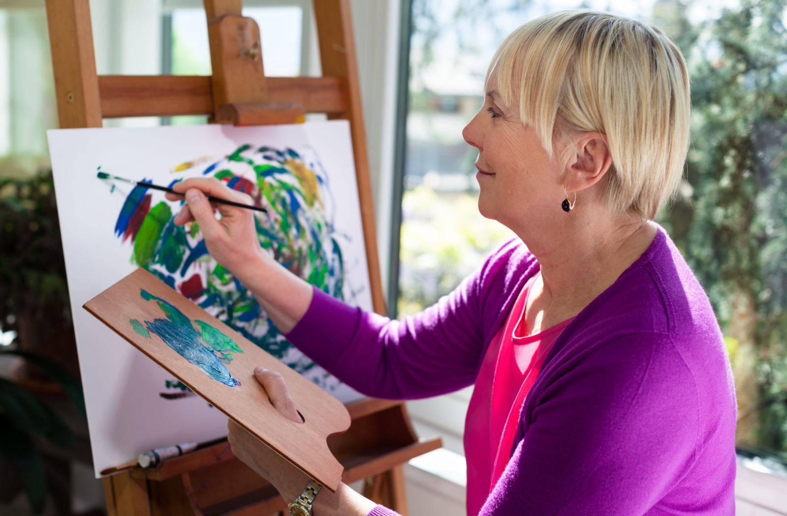 An older woman painting on a canvas with a palette in hand, in a sunlit room.