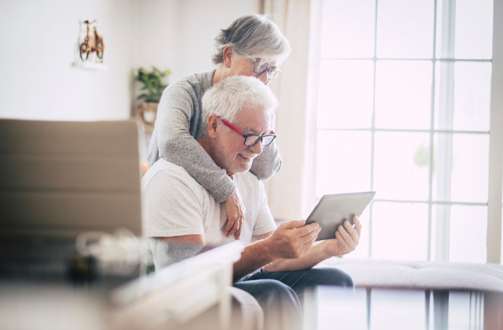 An older couple laughing in their sunlit kitchen while looking at photos of an assisted living community on their tablet.