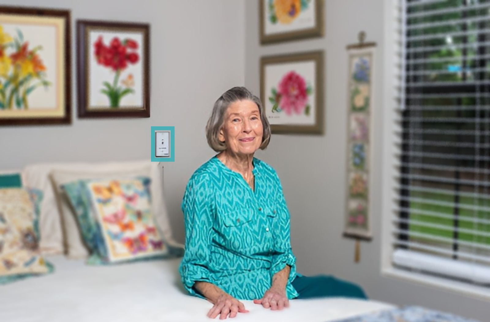 A senior woman sitting on her bed with the staff call button highlighted in the back.