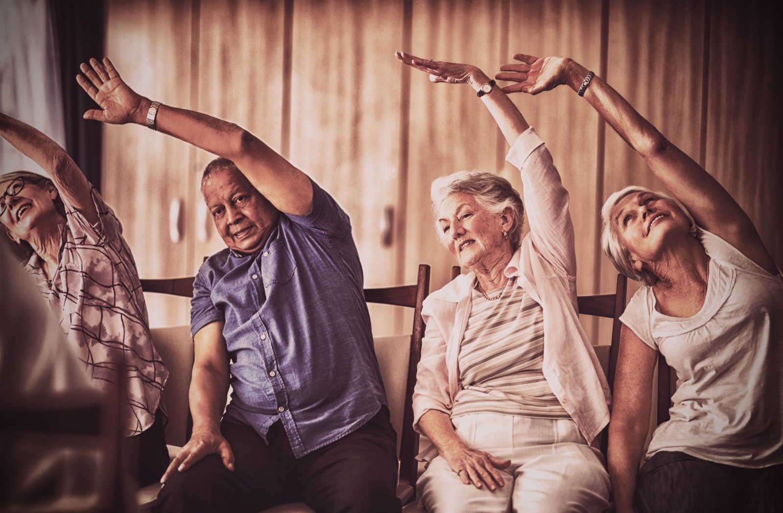 Four seniors lift their hands over their heads in a seated chair stretch, following the movements of an instructor
